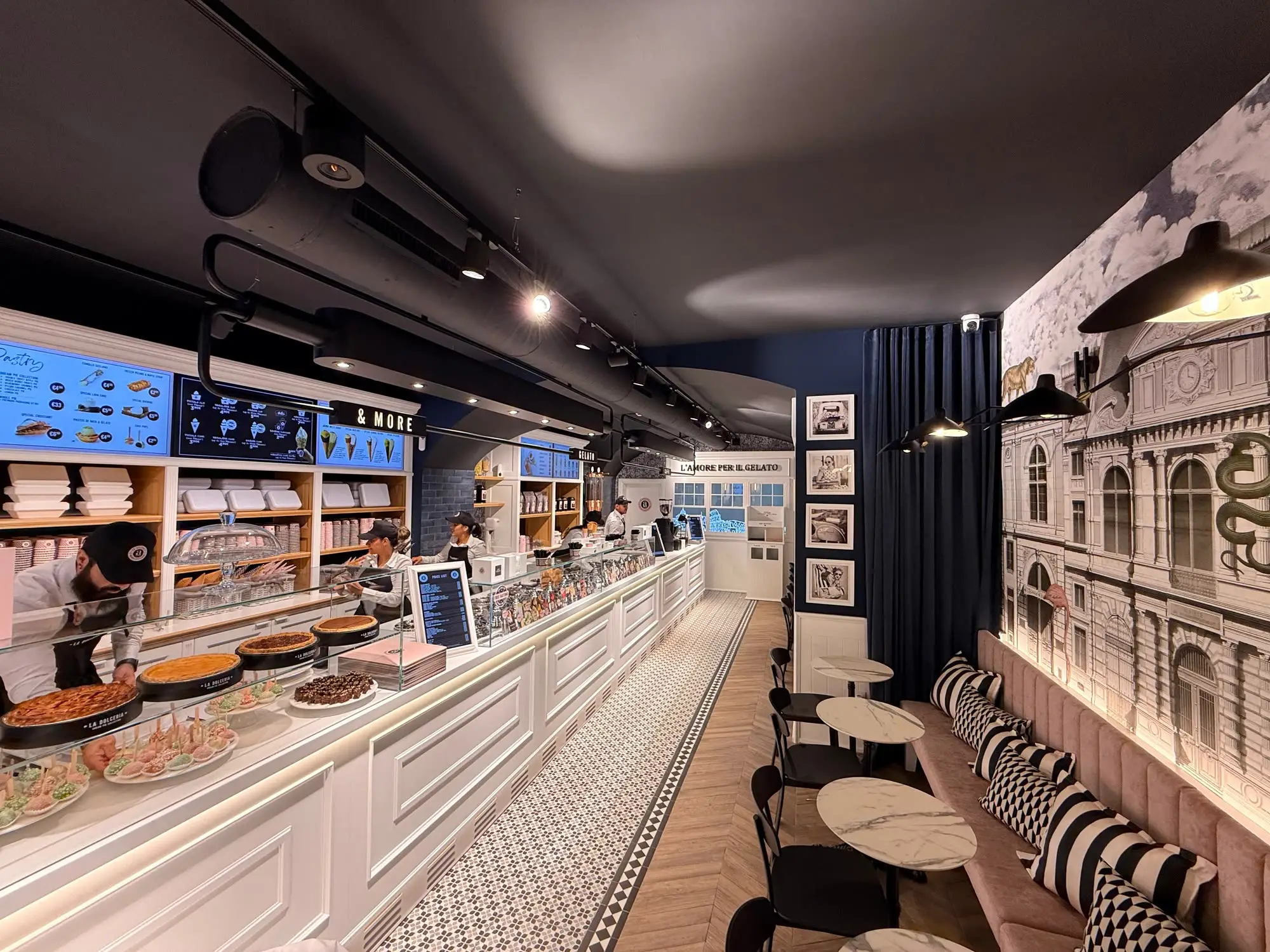 Interior da La Dolceria aROMA: balcão em branco ao longo da sala, tetos abobadados e mural de fachadas lisboetas na parede oposta