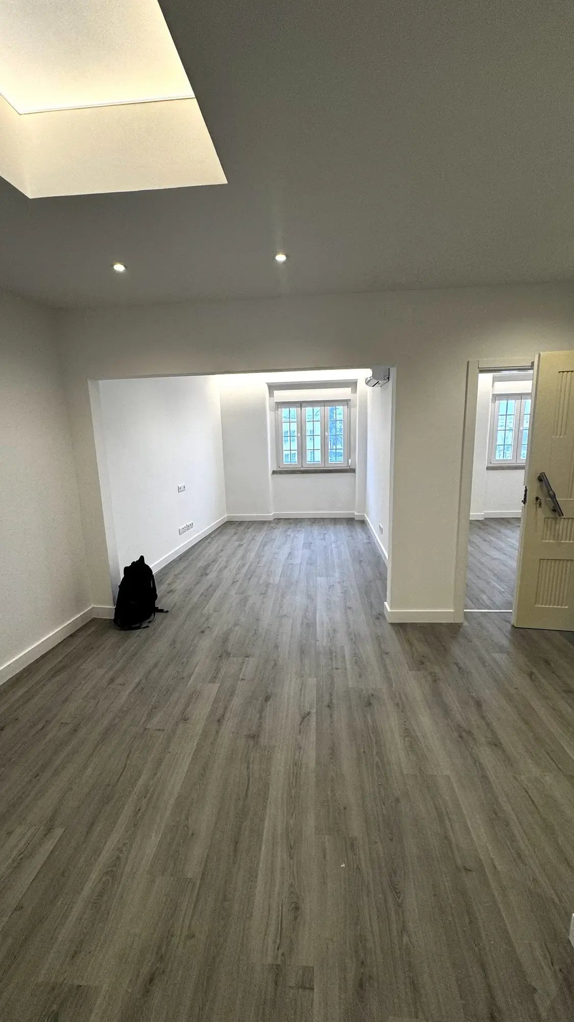 Sala acabada com pavimento vinílico cinzento, janela ao fundo, ar condicionado instalado e a porta de entrada aberta para o corredor
